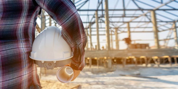 "Un trabajador de la construcción con casco y plano frente a una estructura en construcción."